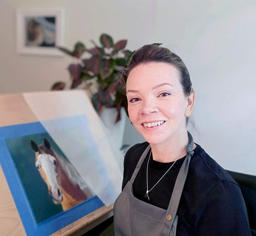 Kristen Ohlsson sitting in studio next to a realistic pastel drawing of a horse.